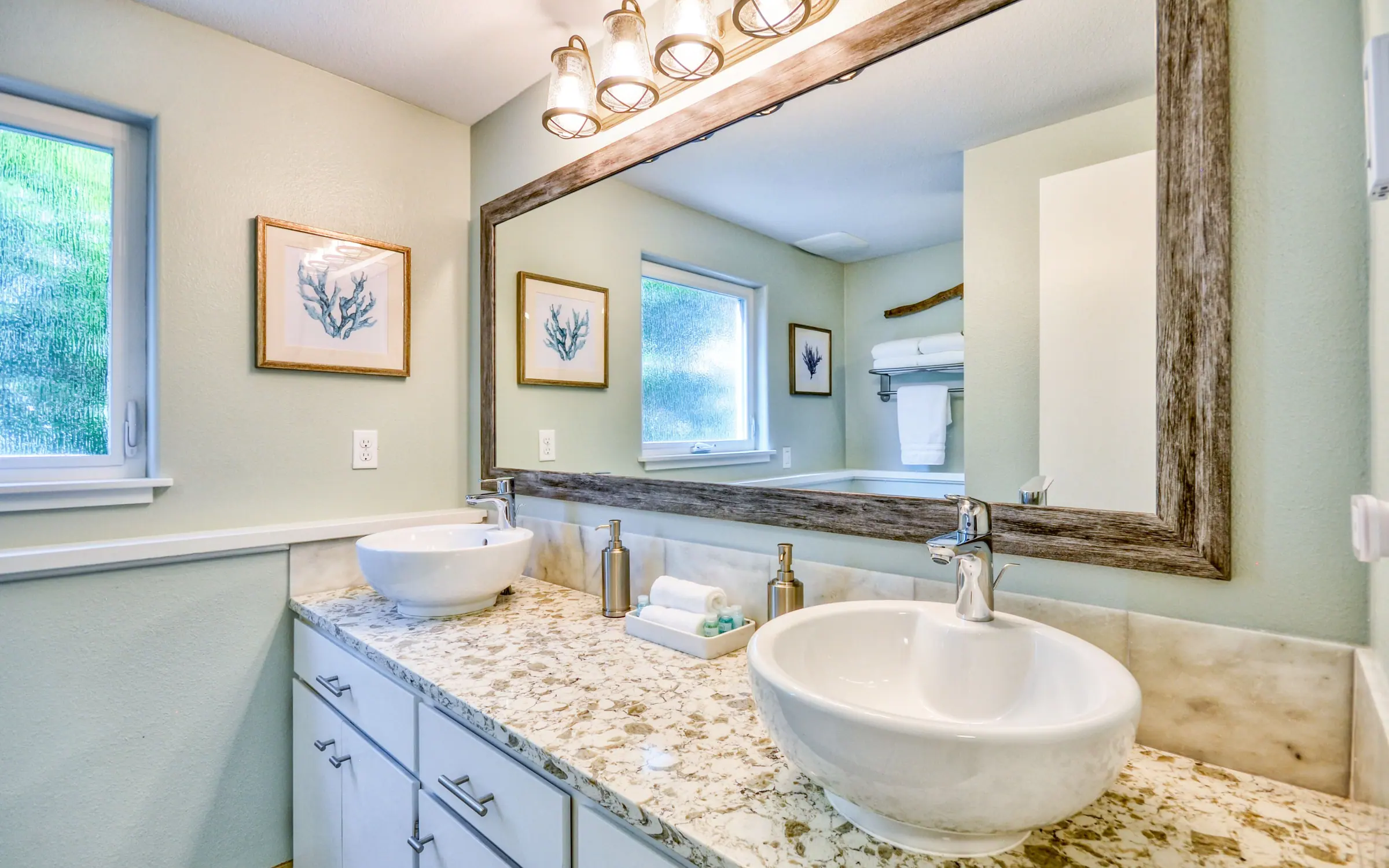 A bathroom with two white vessel basins on a speckled stone counter, a wide mirror in a pale driftwood frame, framed coastal prints on the walls, and folded white towels on a rail.