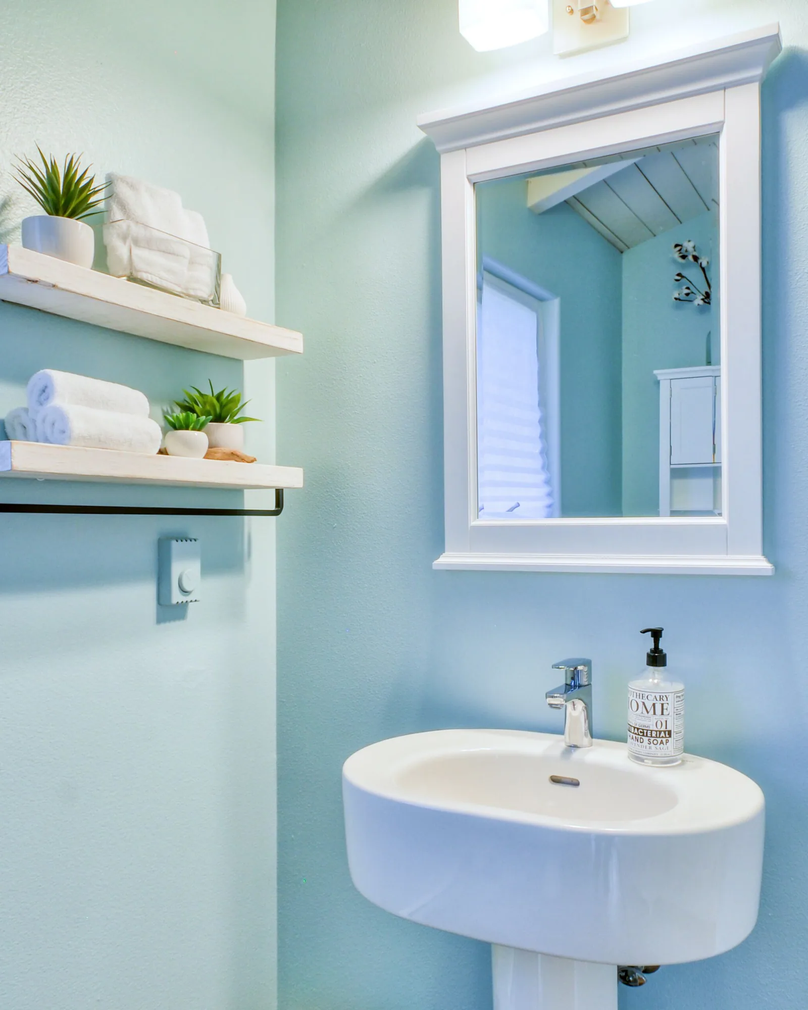 The second bathroom: a white pedestal basin against pale blue walls, a framed mirror above it, an open shelf with rolled towels and a small plant, and a shuttered window to the left.