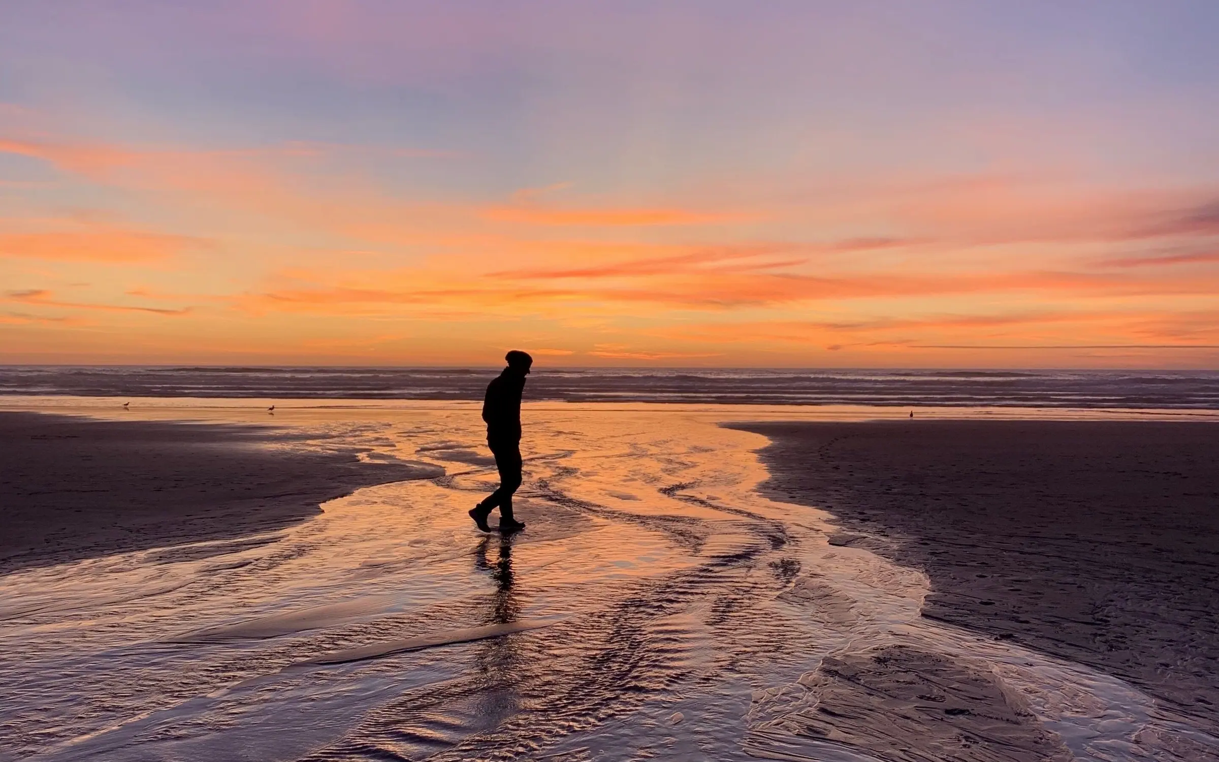 A single figure walking across wet sand at sunset, their reflection and the orange sky mirrored in the shallow water, with a low band of cloud along the horizon.