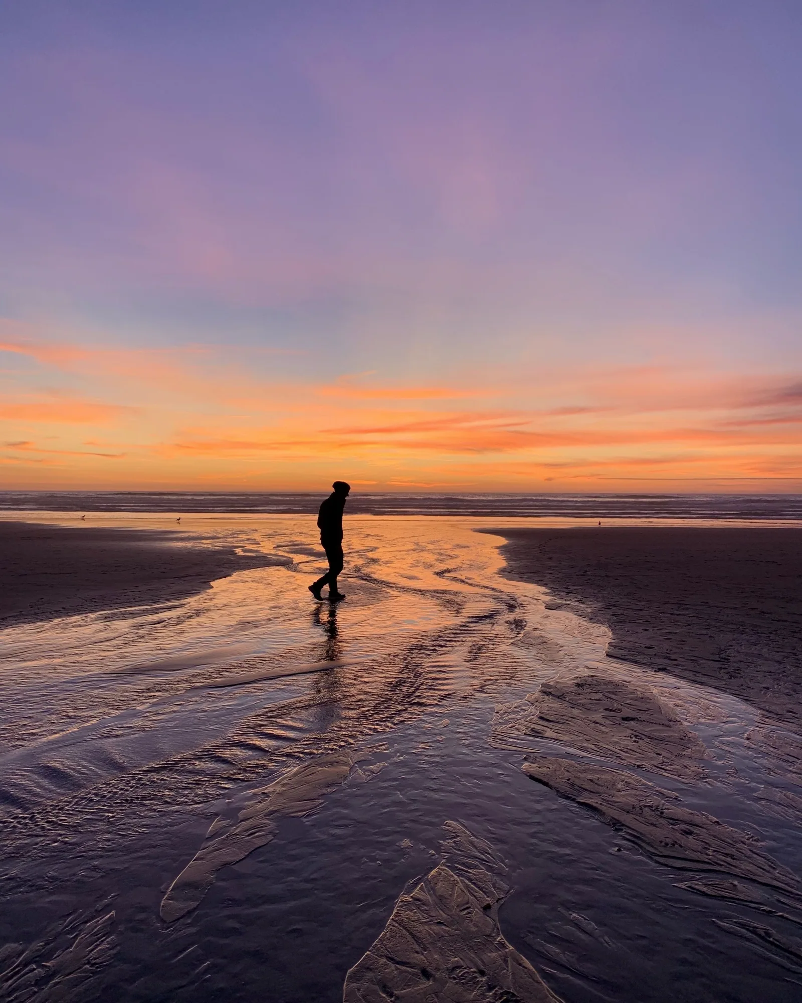 A single figure walking across wet sand at sunset, their reflection and the orange sky mirrored in the shallow water, with a low band of cloud along the horizon.