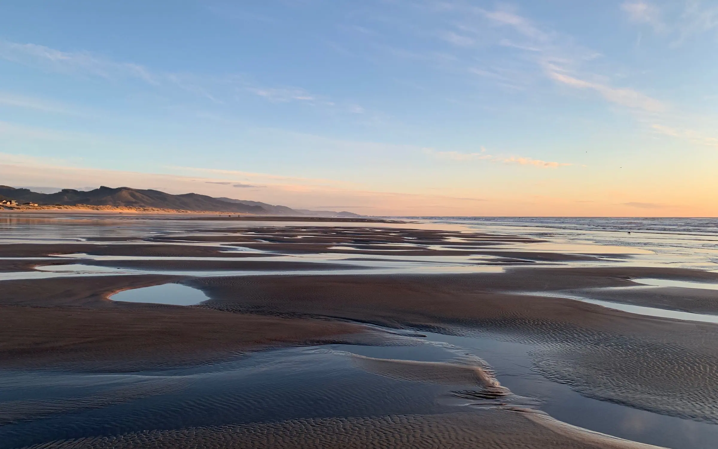 Shallow pools left across a wide flat beach at low tide, mirroring a soft pink and blue sky, with the dune line and the town’s treeline low on the left horizon.