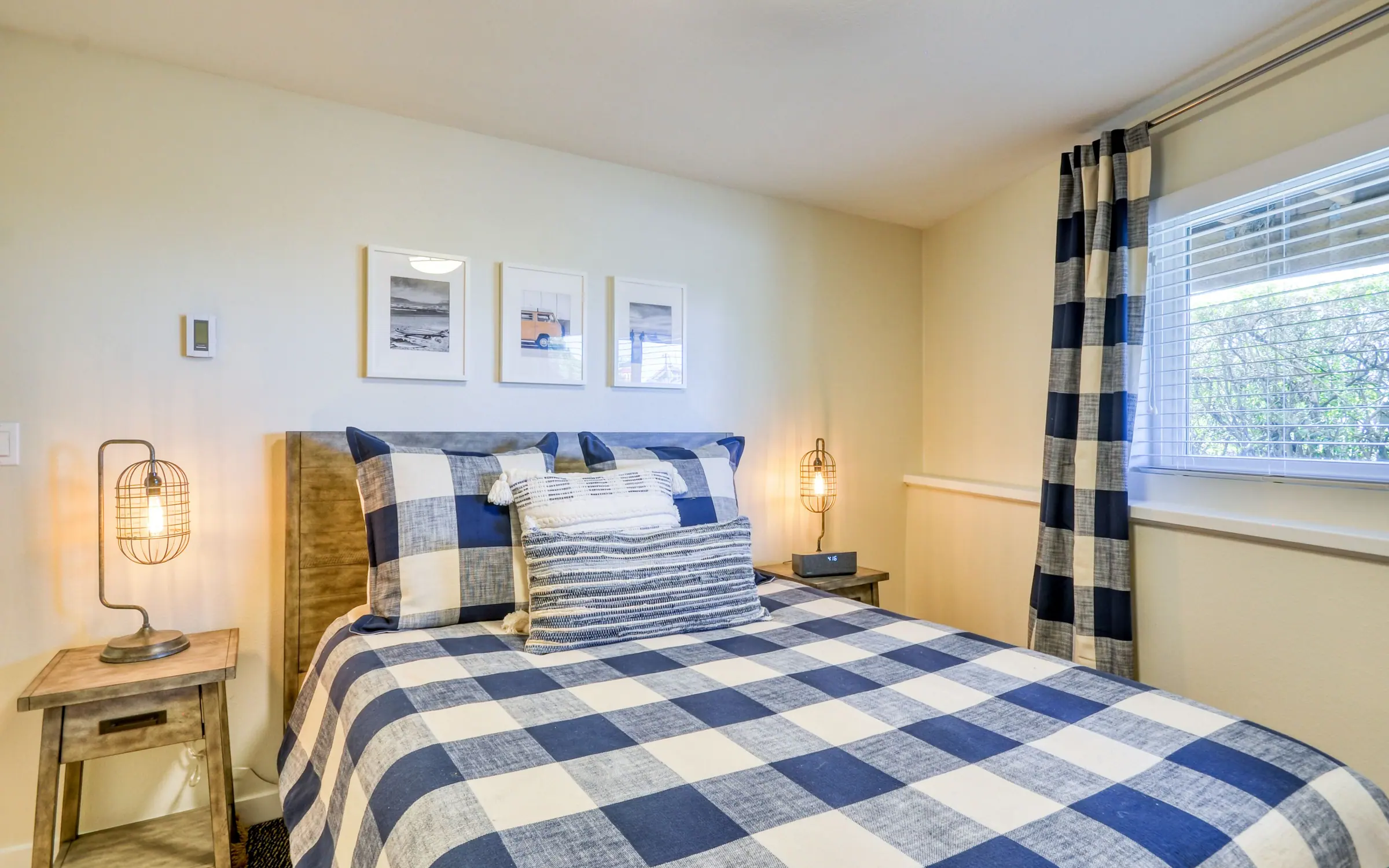 Riptide: a double bedroom with navy and white buffalo-check bedding, a woven headboard, matching bedside lamps and framed prints, and a curtained window looking into shore pine.