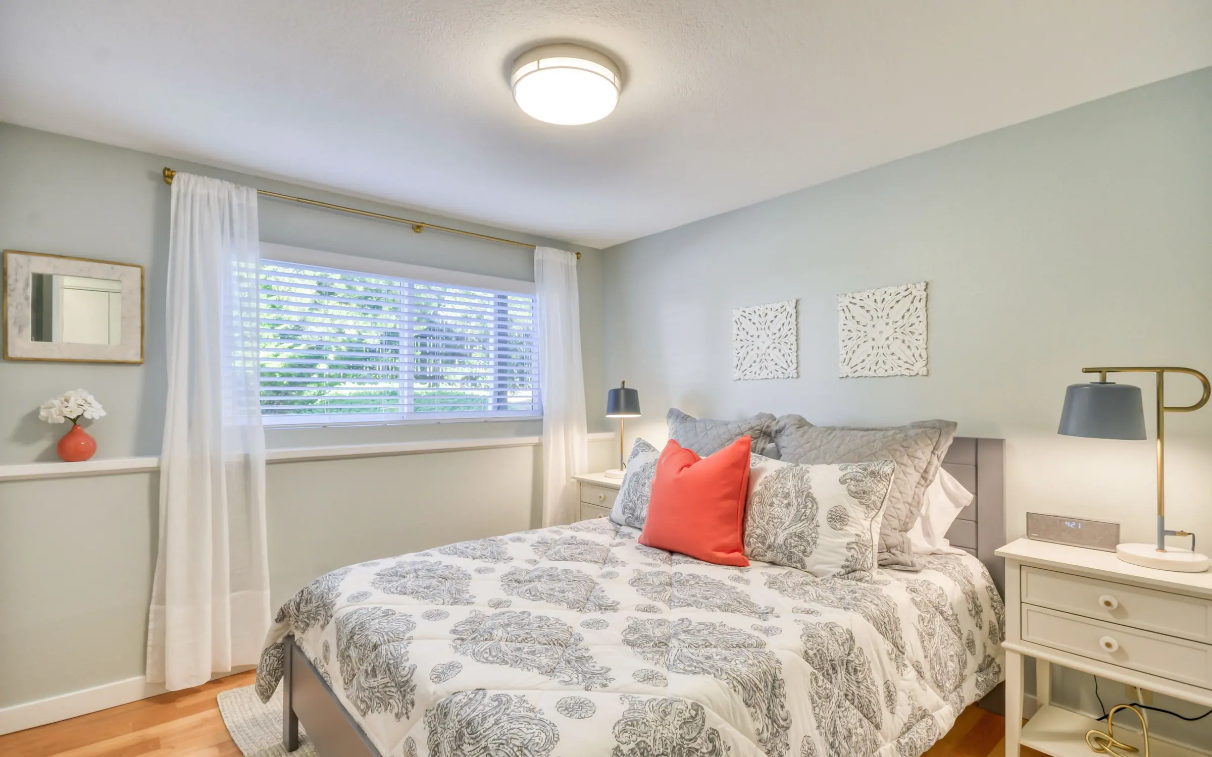 Athena: a queen bedroom with a gray and white patterned quilt and a coral cushion, a pale wooden bed frame, twin bedside tables with dark lamps, and a curtained window at the far end.