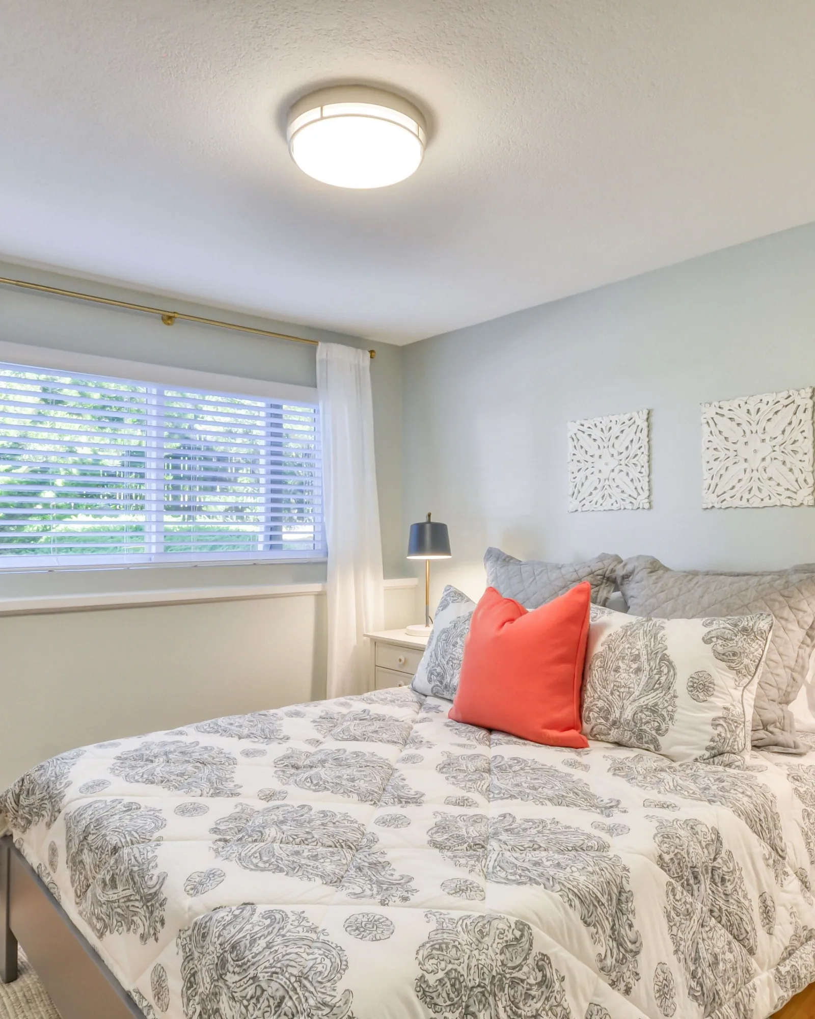 Athena: a queen bedroom with a gray and white patterned quilt and a coral cushion, a pale wooden bed frame, twin bedside tables with dark lamps, and a curtained window at the far end.