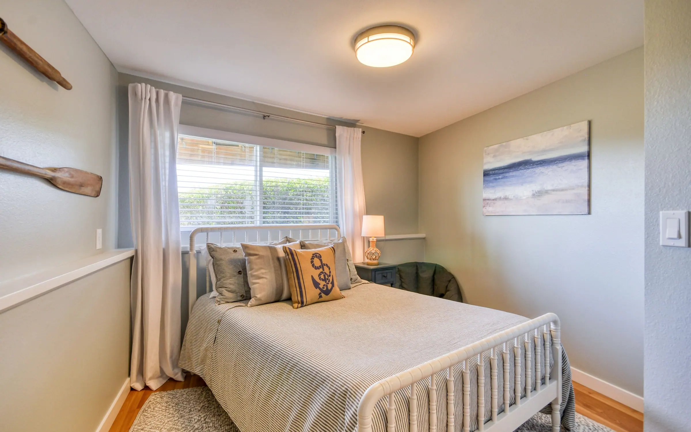 A bedroom with a metal-framed bed, striped and patterned cushions, a driftwood sculpture over the headboard, a framed beach print, and a curtained window letting in soft daylight.