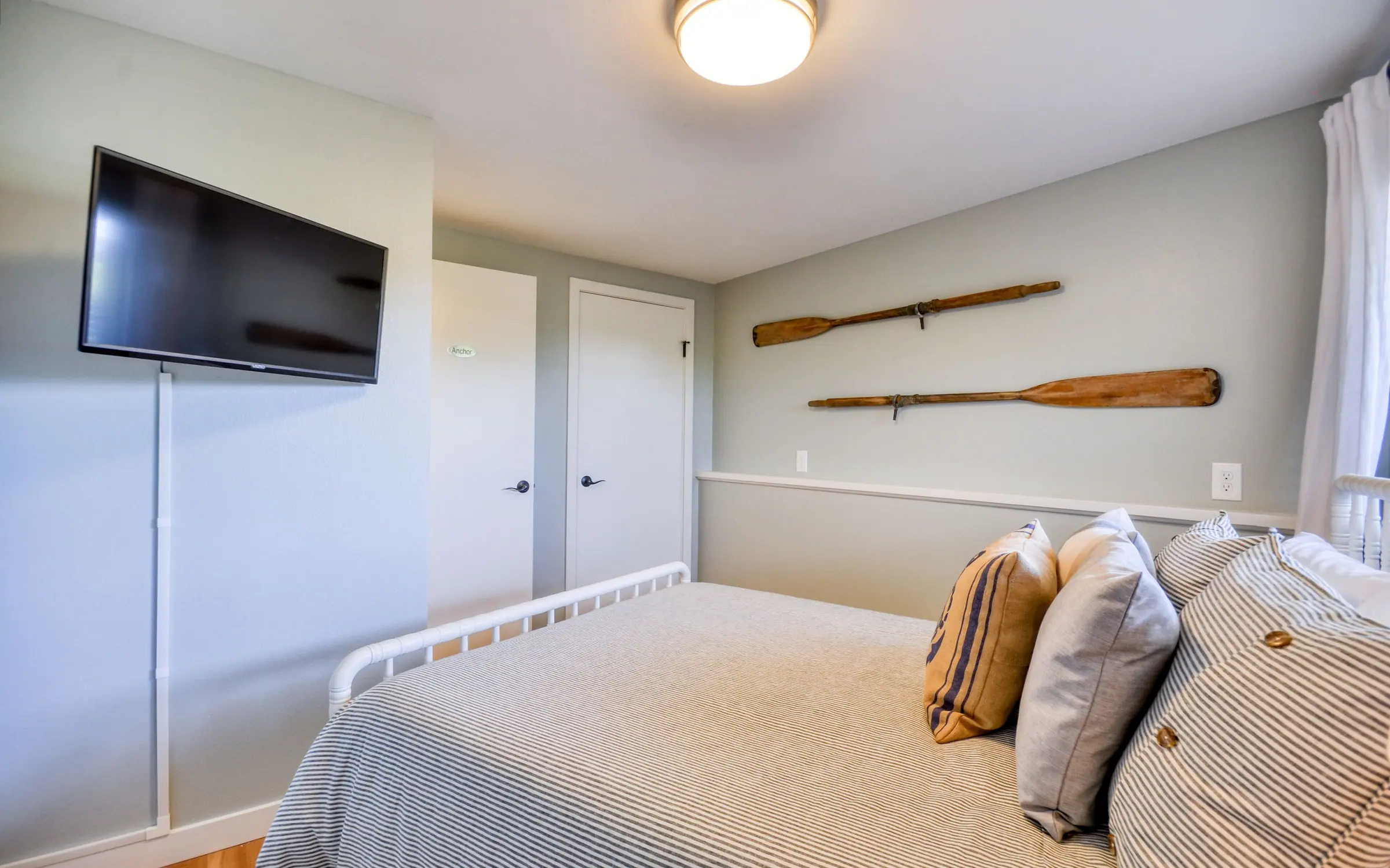 The Anchor room: a bed with striped and woven cushions against a pale wall, a wall-mounted television opposite, a pair of crossed wooden oars hung as art, and a built-in cupboard alongside.
