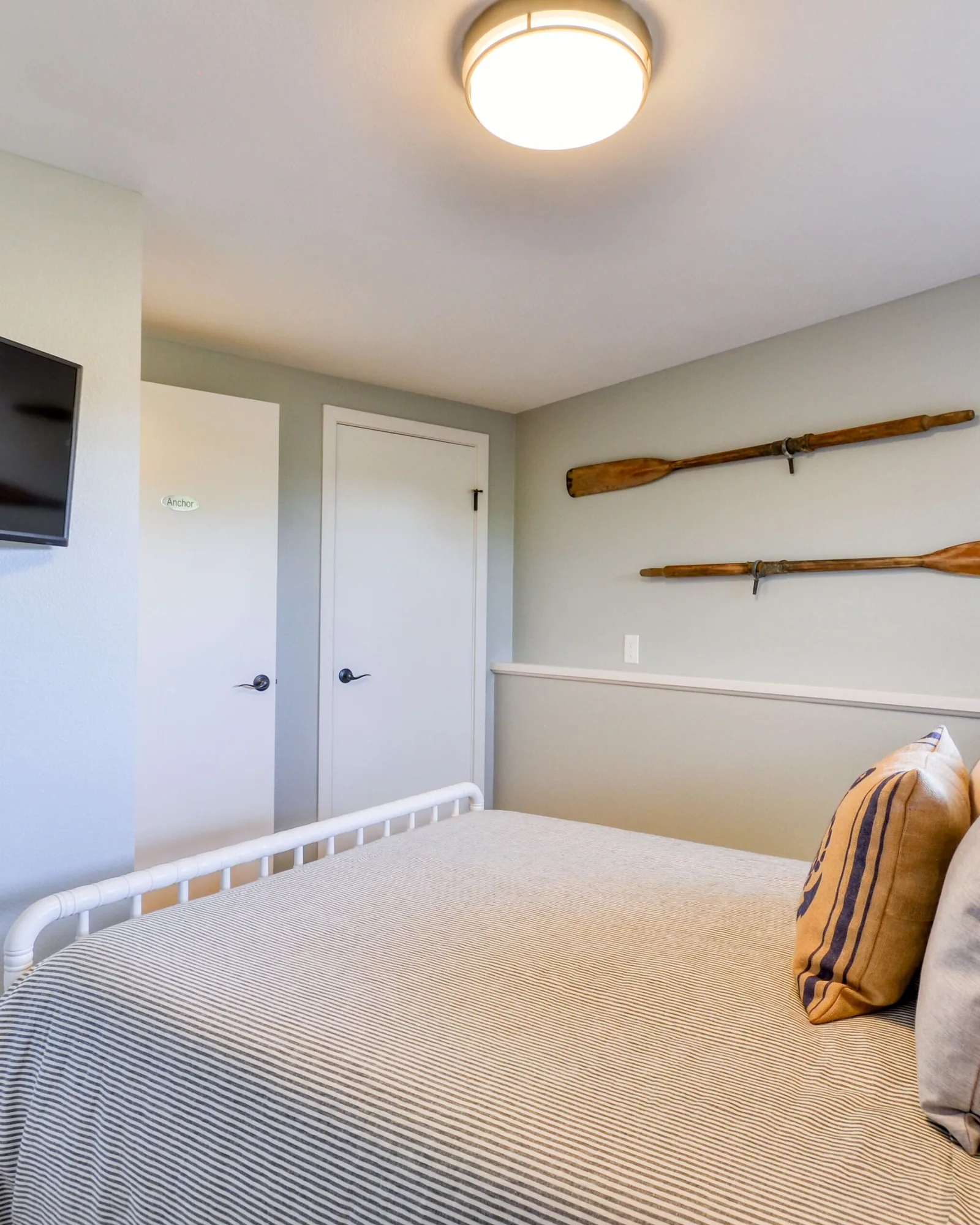 The Anchor room: a bed with striped and woven cushions against a pale wall, a wall-mounted television opposite, a pair of crossed wooden oars hung as art, and a built-in cupboard alongside.