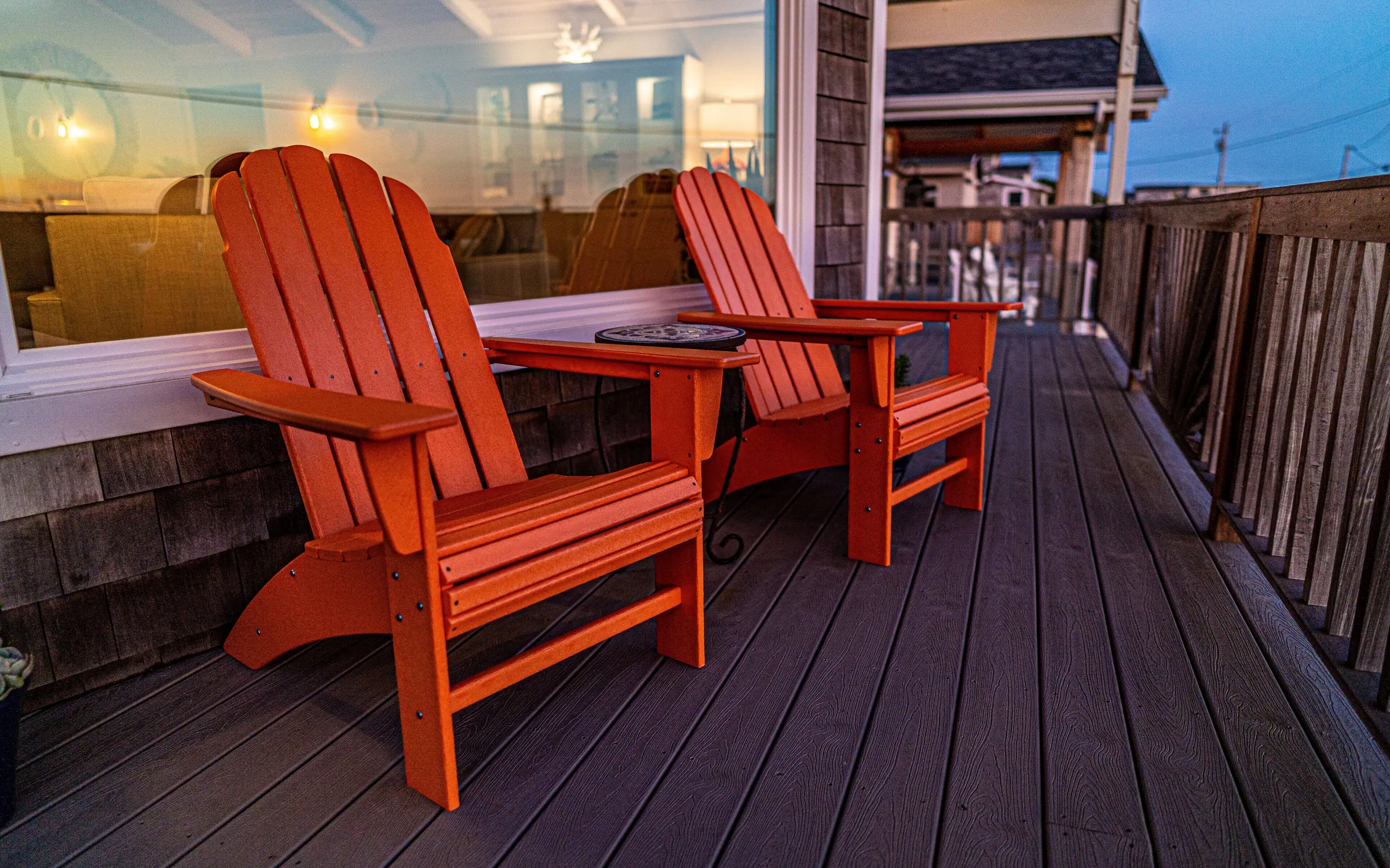 Two orange Adirondack chairs with a small round table between them on the composite deck at dusk, warm light spilling from the great-room windows behind, and the neighboring rooflines and power lines beyond the rail.