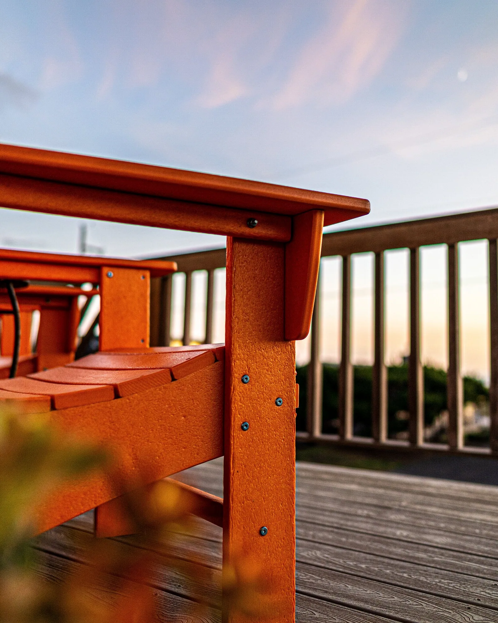 Looking along the deck rail at sunset past the arm of a wooden Adirondack chair, the sky banded orange and blue above the balusters.