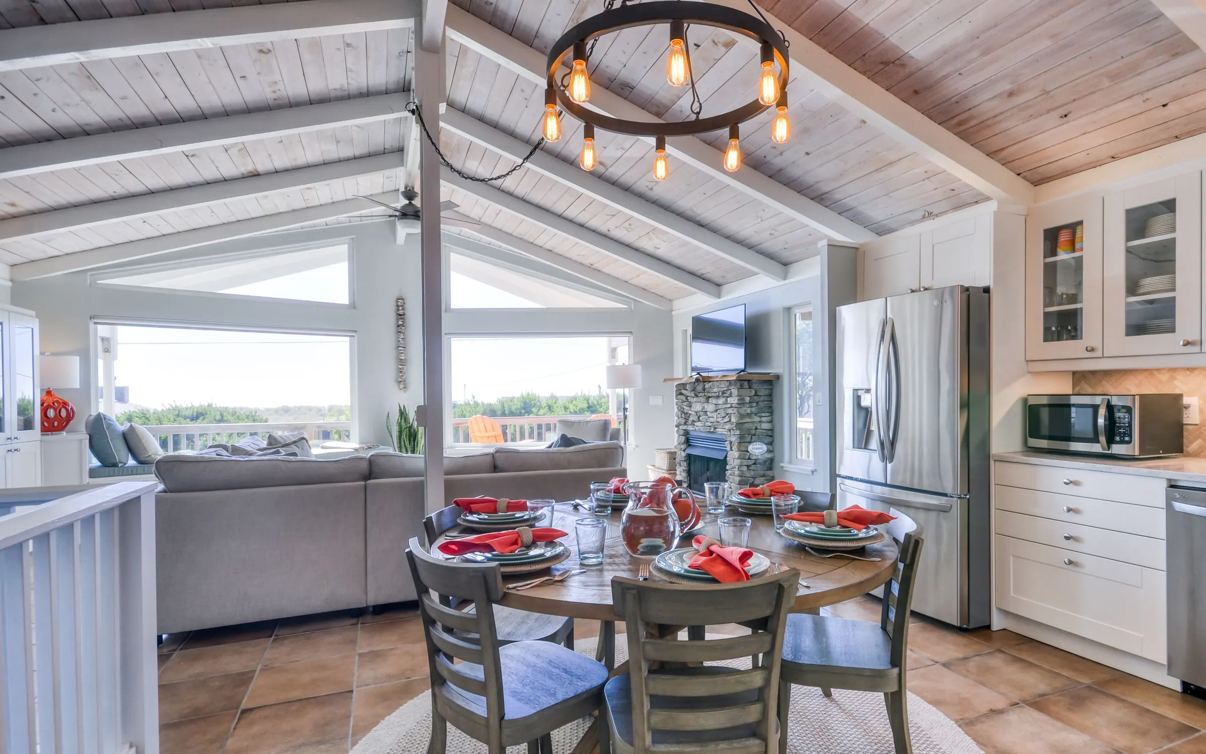 A round wooden dining table laid for six with coral napkins and a water pitcher, the stacked-stone fireplace and the sectional behind it, and the kitchen with its stainless refrigerator to the right.