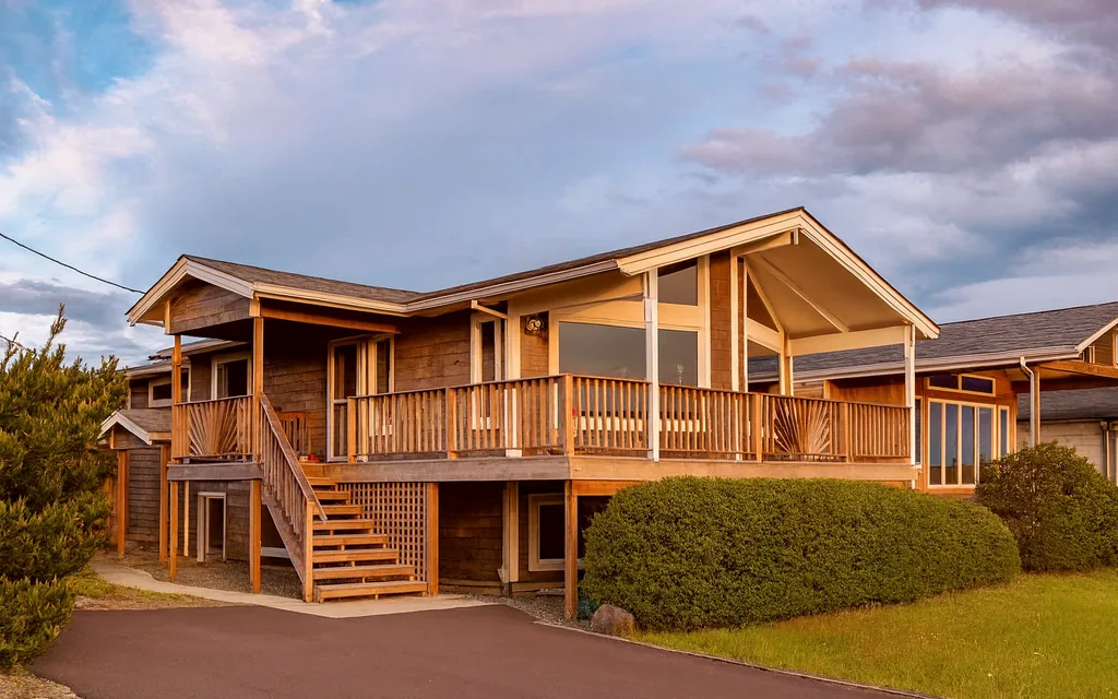 The front of Nostos at sunset: a cedar-shingled beach house with a wide second-story deck, an open-gabled roof over tall picture windows, and an exterior staircase down to a paved driveway edged with shore pine and clipped hedge.