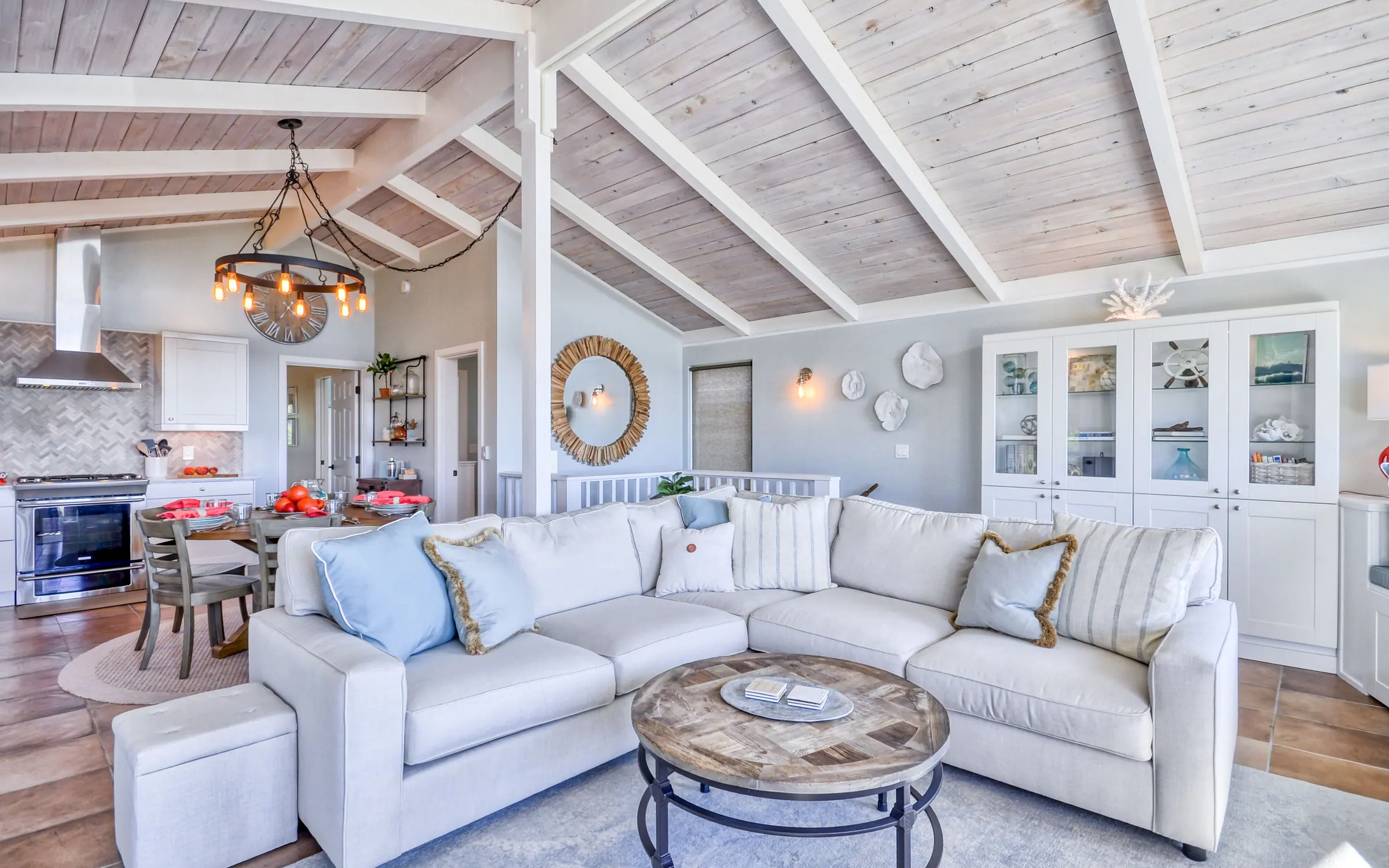 The great room under a whitewashed plank cathedral ceiling with white beams: a large pale linen sectional around a round wood coffee table, an iron ring chandelier, and the kitchen and dining table visible beyond.