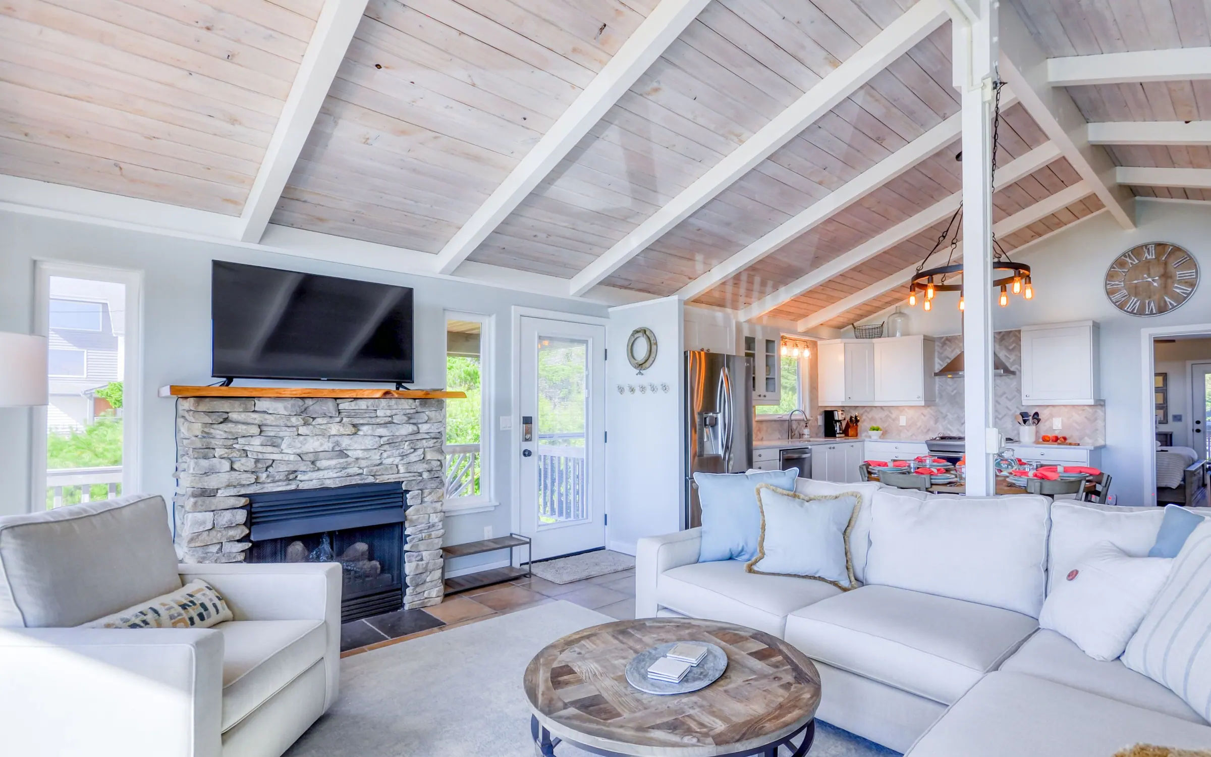 The stacked-stone fireplace with a live-edge wood mantel and a wall-mounted television above it, a glazed door to the deck alongside, and the kitchen and dining table further down the open plan.