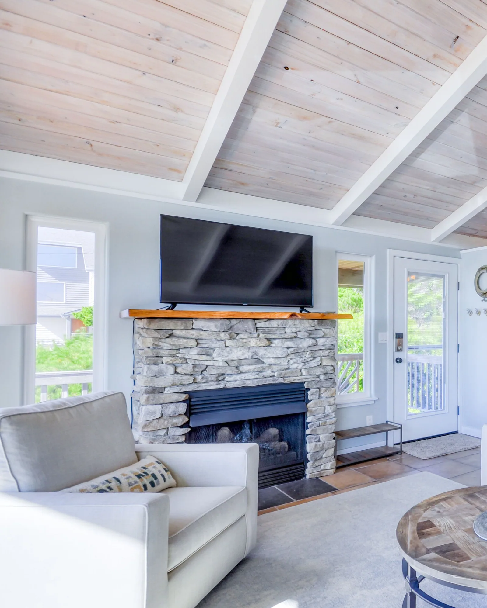 The stacked-stone fireplace with a live-edge wood mantel and a wall-mounted television above it, a glazed door to the deck alongside, and the kitchen and dining table further down the open plan.