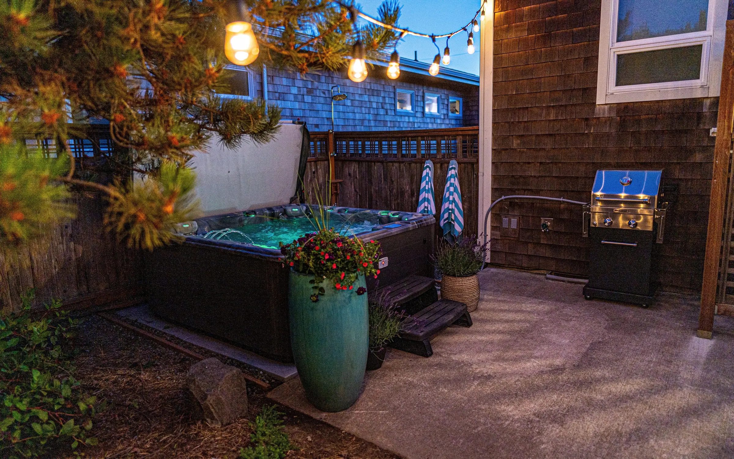 The hot tub at dusk with its cover propped open and the water lit turquoise, festoon bulbs strung overhead through a shore pine, two striped towels hung ready, and a propane grill on the concrete pad alongside.