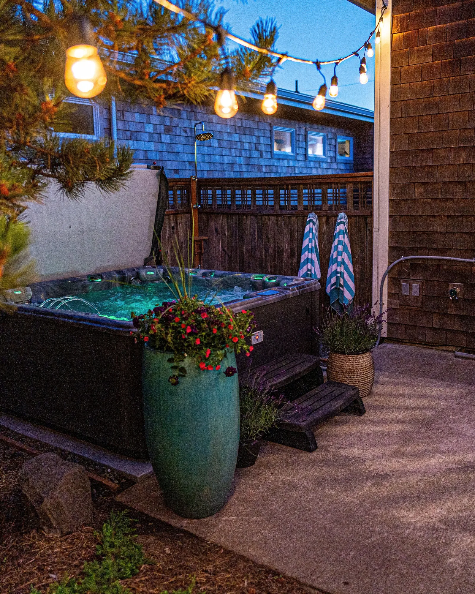 The hot tub at dusk with its cover propped open and the water lit turquoise, festoon bulbs strung overhead through a shore pine, two striped towels hung ready, and a propane grill on the concrete pad alongside.