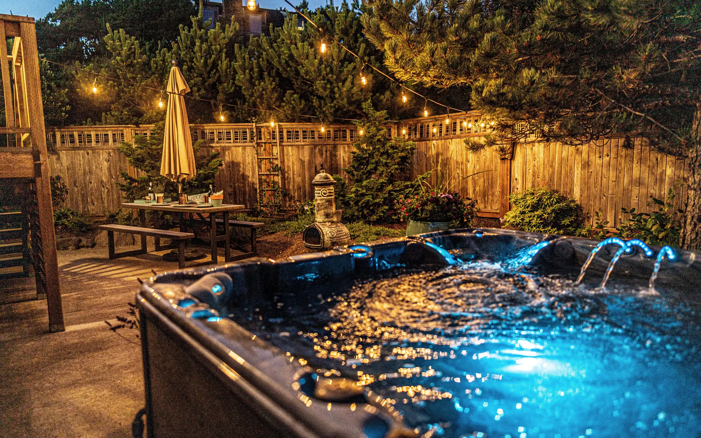 The hot tub after dark, water glowing blue, ringed by festoon lights along the cedar fence, with a folded umbrella, a picnic table and planted pots in the enclosed backyard behind it.