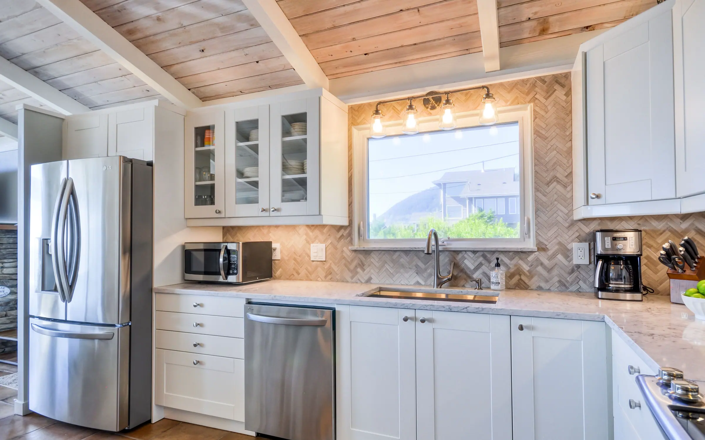 The working side of the kitchen: a deep sink under a window that looks out to shore pine, dishwasher below the counter, coffee maker and grinder on the worktop, and a tall stainless french-door refrigerator at the end of the run.