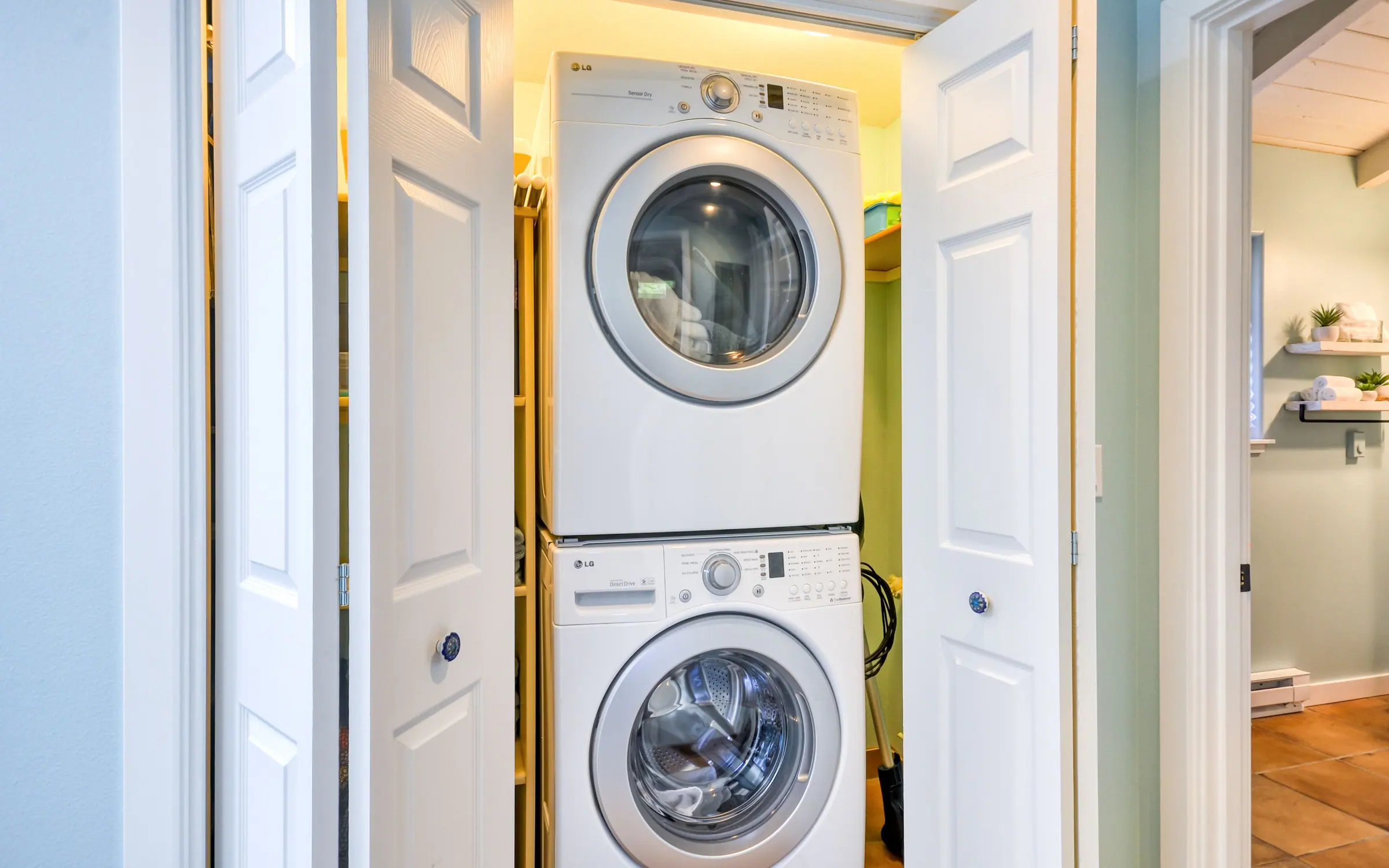 A stacked white front-loading washer and dryer in a lit closet with louvered doors open, alongside the pale hallway of the house.