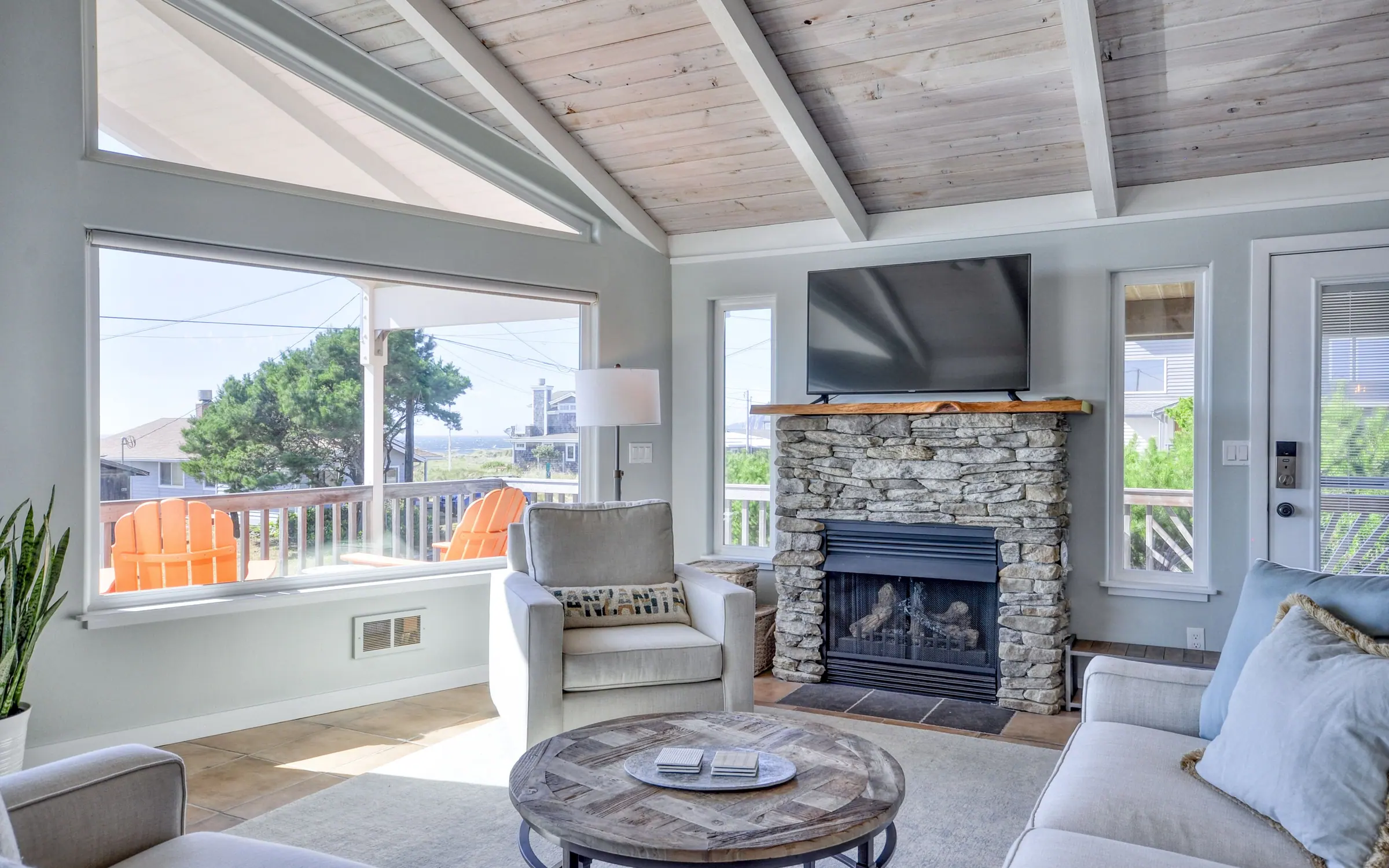 The living end of the great room: a stacked-stone fireplace with a television above it, pale sofas and a round wood coffee table on a light rug, and daylight coming in from the picture windows on the right.