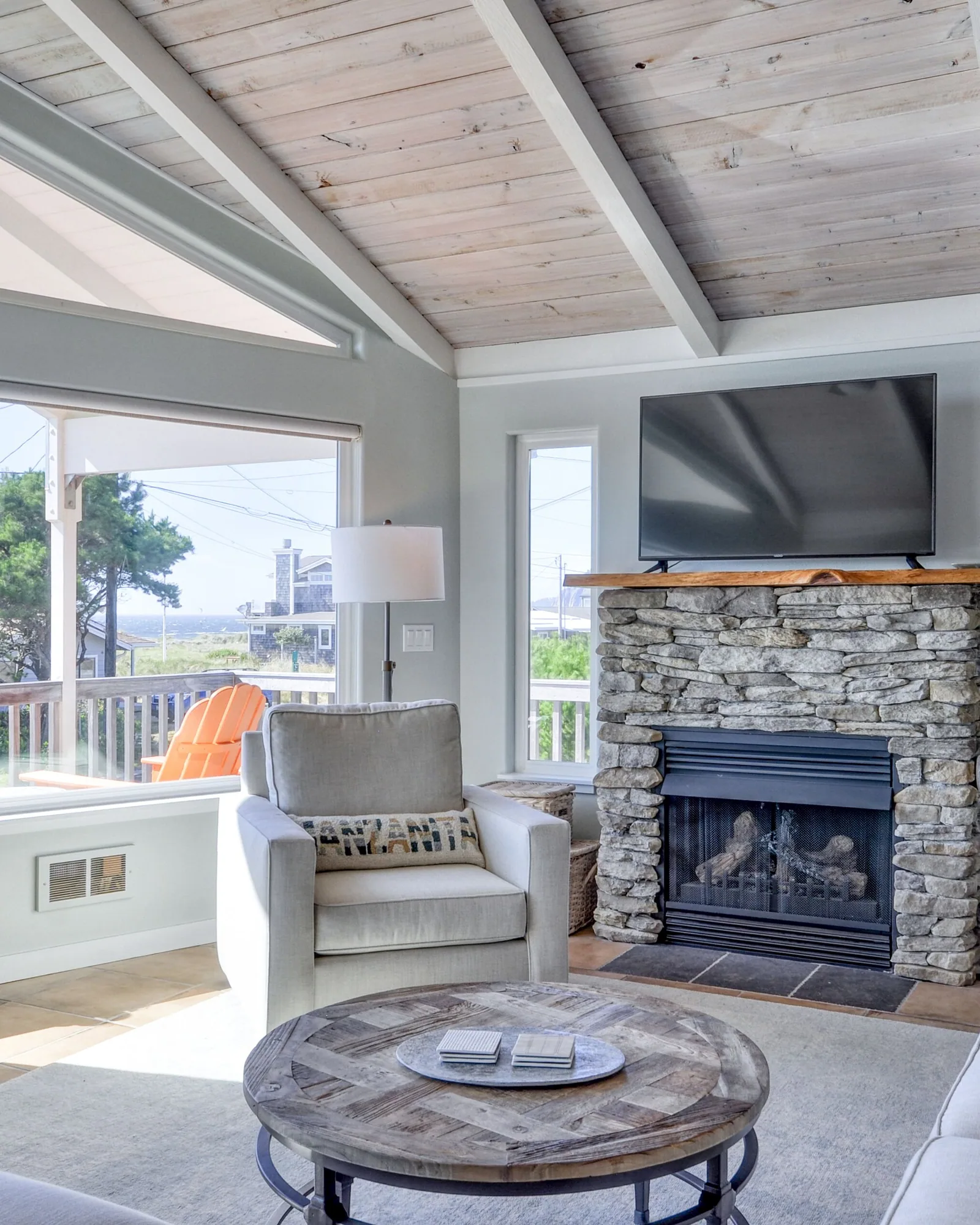 The living end of the great room: a stacked-stone fireplace with a television above it, pale sofas and a round wood coffee table on a light rug, and daylight coming in from the picture windows on the right.