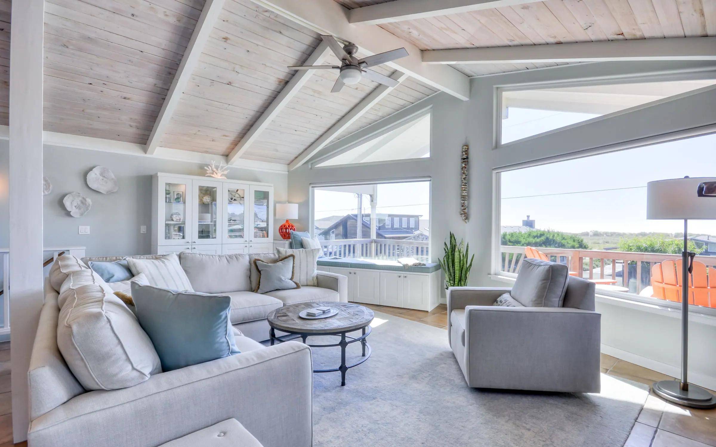 The seating end of the great room looking out through tall picture windows and a triangular gable window to the deck, where orange Adirondack chairs face the dune grass. A built-in window seat runs beneath the glass.