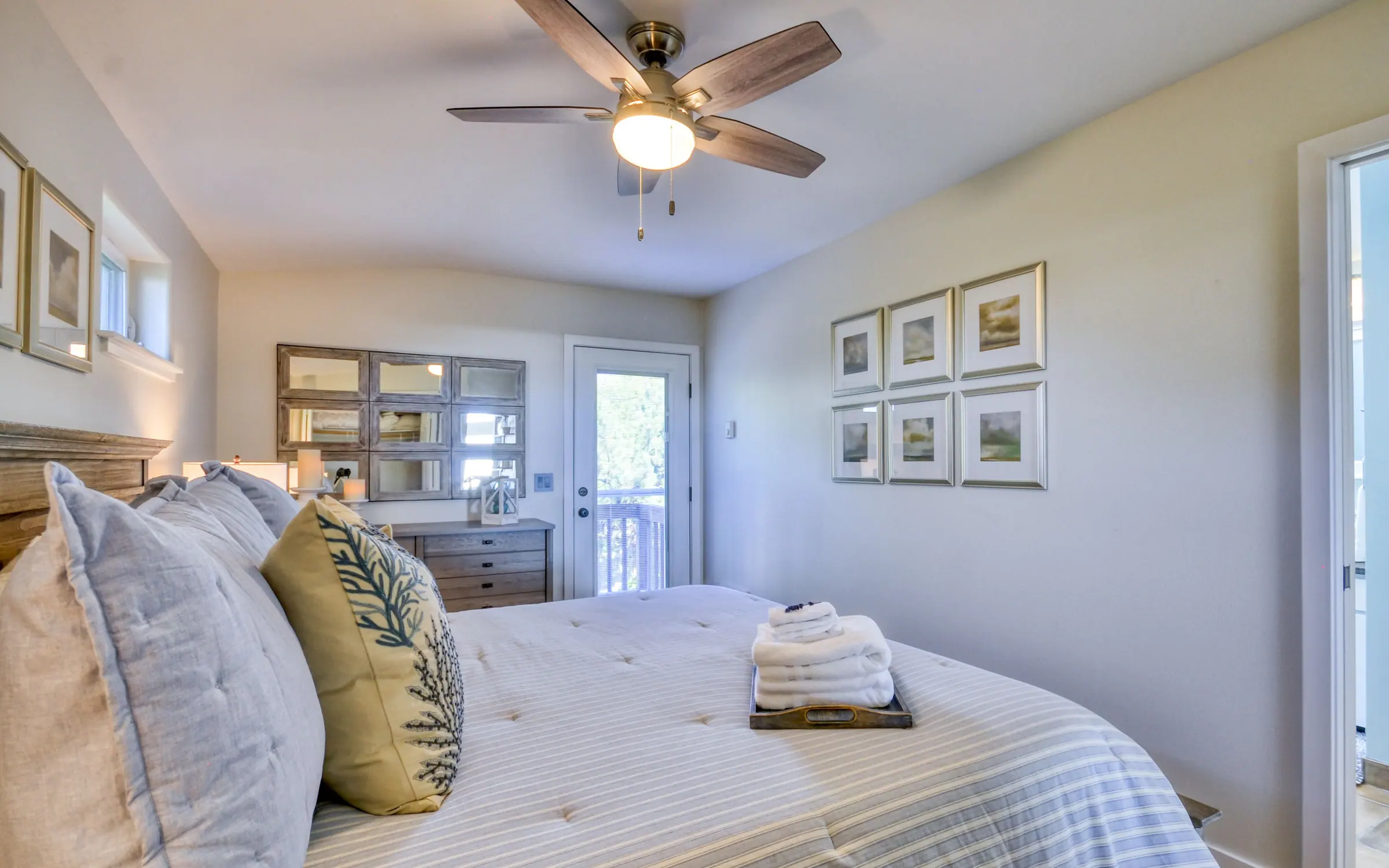 The Master Suite: a king bed with a wooden headboard and blue-gray striped linen, a row of framed prints above it, a ceiling fan, a wide dresser, and a glazed door out to the deck.
