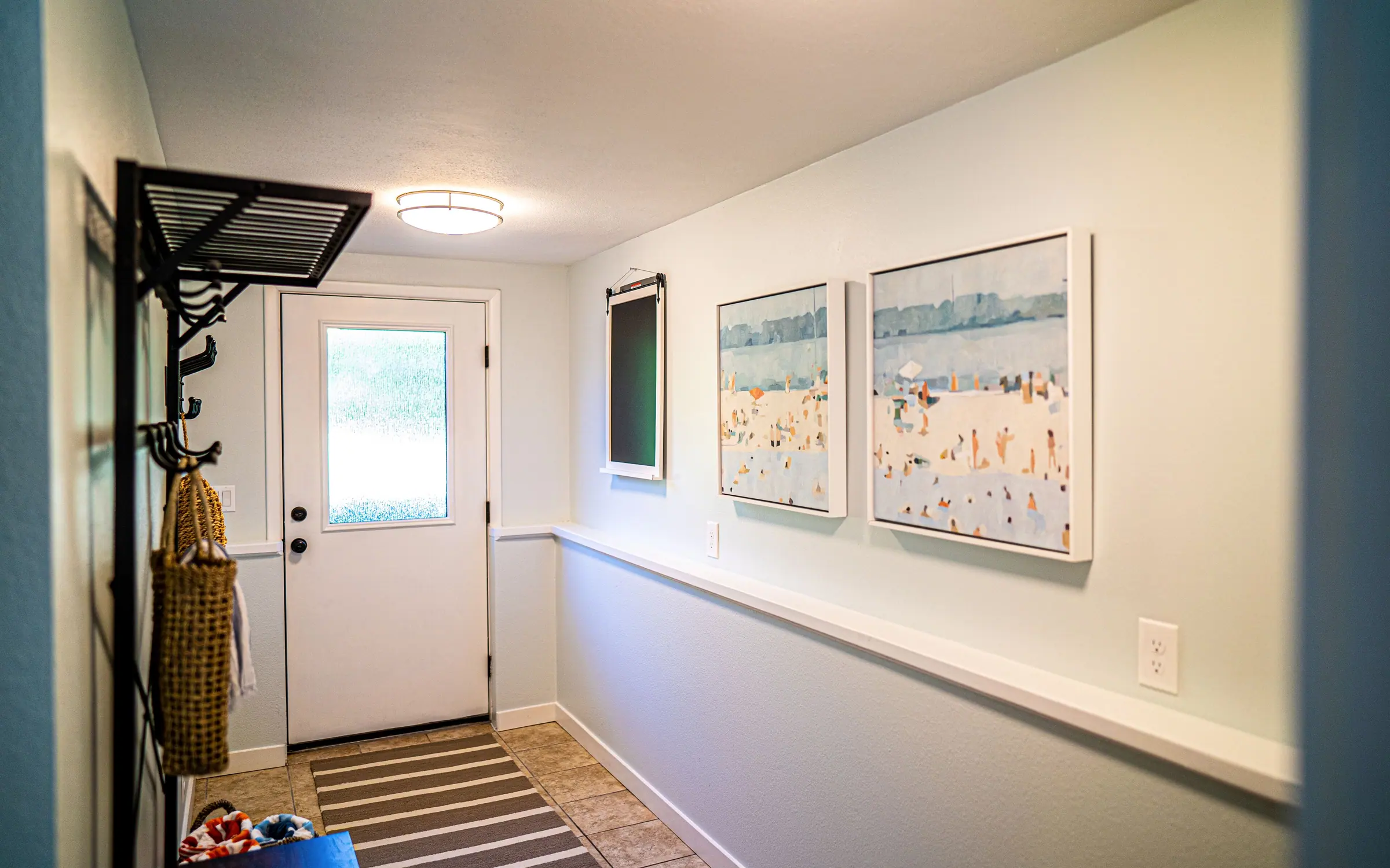 The mud room and entry hall: a white door with a window, a wall rack of coat hooks with a bag hanging from it, two framed beach prints, and a runner over the floor leading into the house.