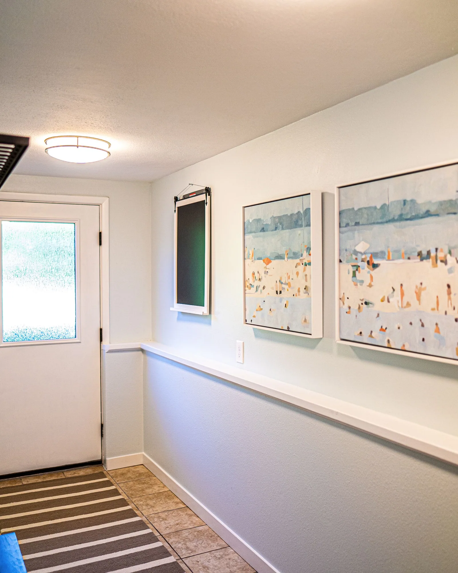 The mud room and entry hall: a white door with a window, a wall rack of coat hooks with a bag hanging from it, two framed beach prints, and a runner over the floor leading into the house.