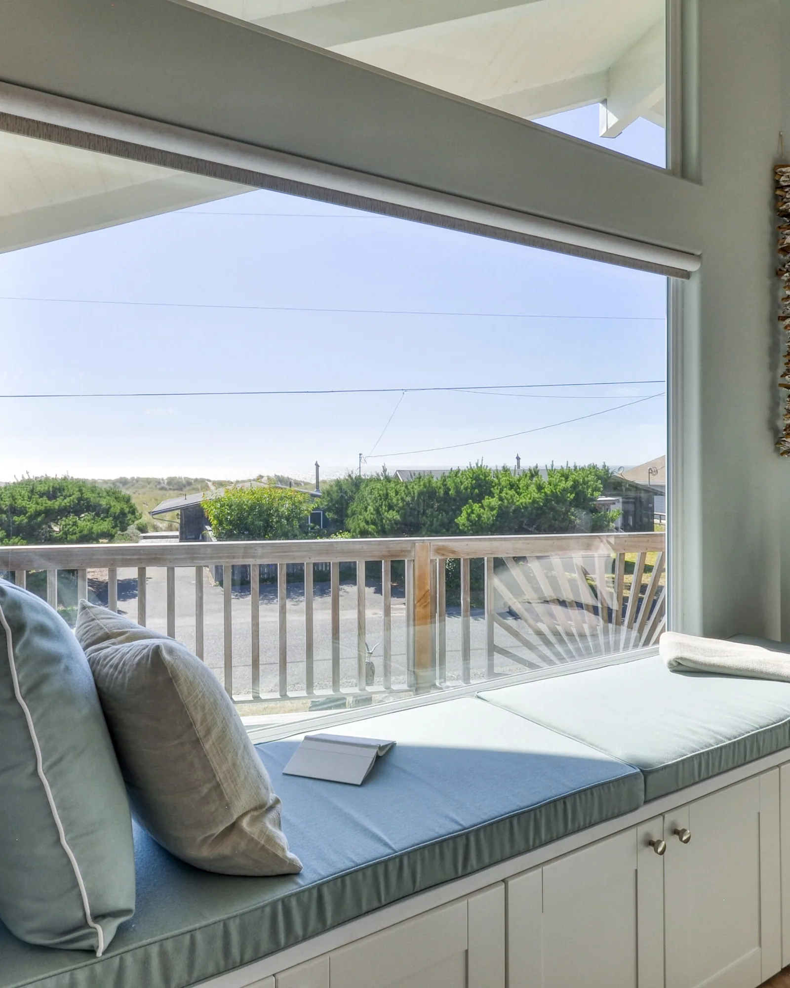 A built-in window seat with pale green cushions and two pillows, set beneath a wide picture window that looks out over the deck railing to the dunes, with cupboard doors below the bench and a potted snake plant beside it.